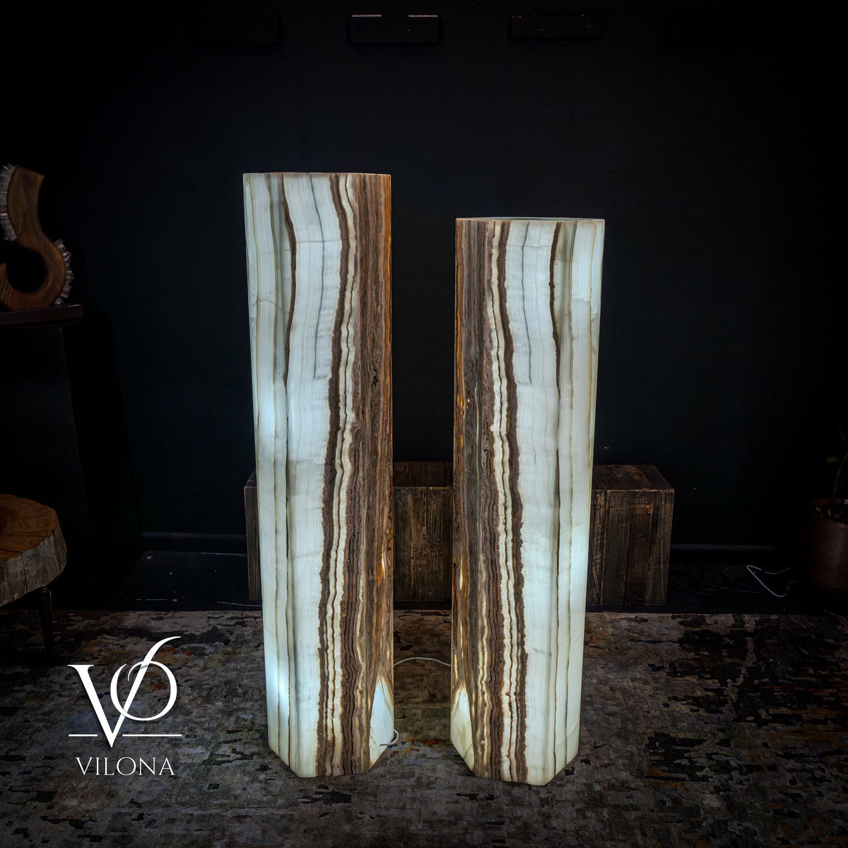 Pair of white onyx floor lamps with vertical brown and beige striations in hexagonal column shapes, standing on a textured rug against a dark wall.