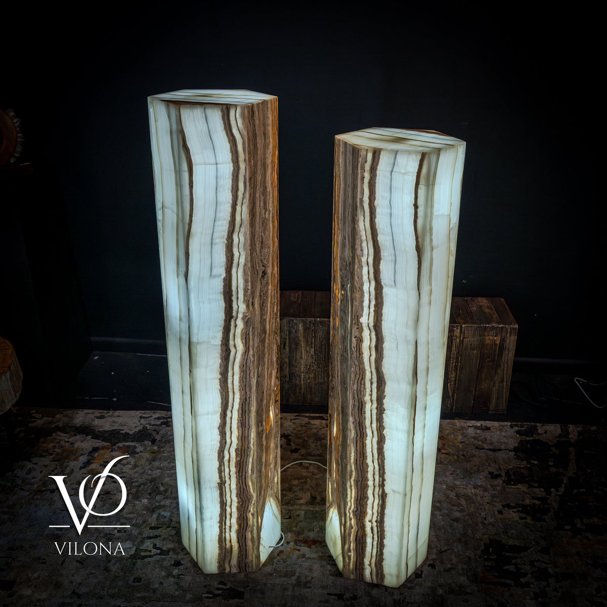 Pair of tall hexagonal floor lamps with translucent white onyx bodies and natural brown banding, softly glowing in a dark room with textured carpet floor.