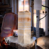Tall rectangular onyx table lamp with creamy white and golden horizontal layers, placed on a rustic wooden slab in a cozy room with natural light and leather chair.