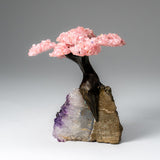 Small ornamental tree sculpture with a dark, twisted trunk and branches holding clustered pink rose quartz crystals, mounted on a rough amethyst-stone base with purple and beige tones.
