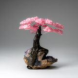 Decorative tree sculpture with clustered pink rose quartz gemstones as leaves and a twisted dark trunk mounted on an amethyst matrix base, photographed on gray background.