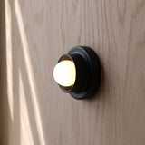 Black metal wall sconce with a rounded, dome-shaped white light fixture mounted on light wood grain wall, showing soft shadows.