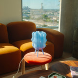 Blue translucent wavy lamp with vertical ridges on three slender legs, casting shadows on an orange cushioned stool in front of a curved orange sofa by a window.