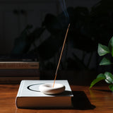 Slim lit incense stick emitting smoke in a smooth white oval holder, resting atop a gray hardcover book on a wooden surface with leafy plant in soft background light.