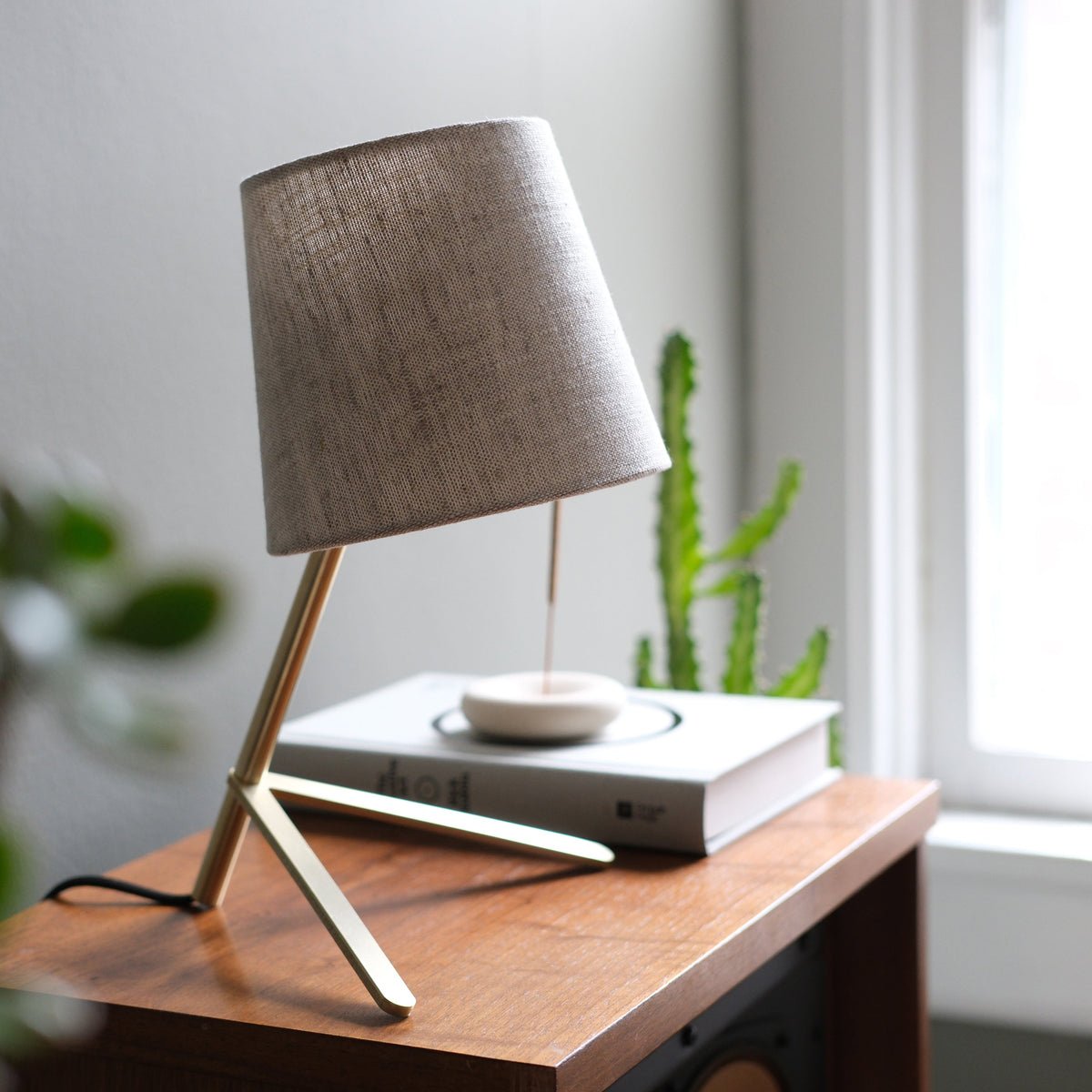 Table lamp with a tilted beige fabric shade and slim, angled metal legs on a wooden side table near a window with a cactus in the background.