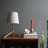 Brass tripod table lamp with beige fabric shade beside stacked book with brass bell topped in red, and a cactus on a wooden side table against gray wall.