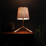 Table lamp with a textured, beige fabric shade and a natural wood tripod base on a wooden surface, softly illuminating nearby small potted cacti against a dark background.