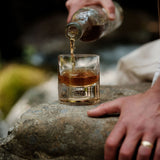 Clear glass tumbler with a metallic bottom detail being filled with amber liquid from a glass bottle, resting on a rough stone surface with a blurred natural background.