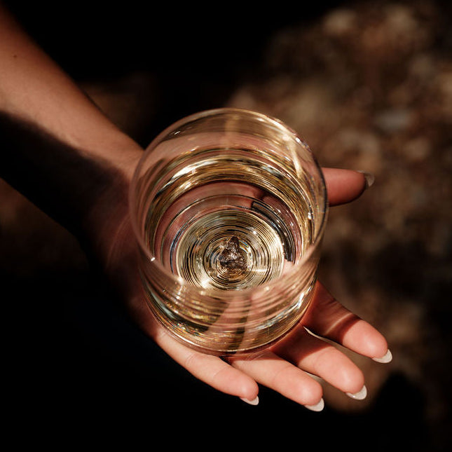 Hand holding a clear glass tumbler viewed from above, showing a swirling pattern and light reflections inside the glass.