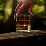 Hand grasping a clear glass tumbler filled with amber liquid, resting on textured wood with a blurred green natural background.