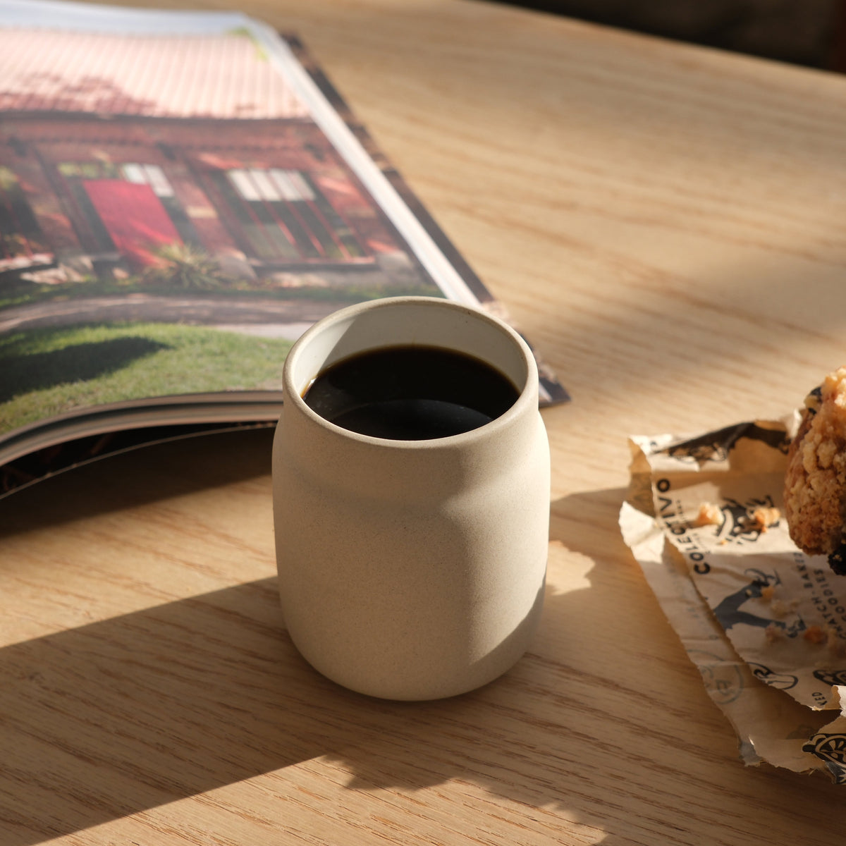 Matte beige ceramic coffee tumbler filled with black coffee on a light wood table, near an open magazine with a photo and a crumpled wrapper with a snack.