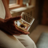 Hand holding a clear glass tumbler with a golden-hued dinosaur bone shard embedded in its base, against a warm, softly lit indoor background.