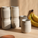 Six beige ceramic mugs with slightly rounded waists arranged on a wooden surface, with a bunch of yellow bananas in the background under warm natural light.
