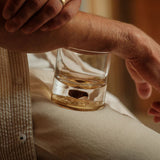 Clear glass tumbler with a brownish dinosaur bone shard embedded at the bottom, resting on a person's beige pants next to their bent arm.