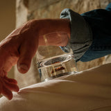 Close-up of a hand holding a clear glass tumbler with a visible embedded shard resembling dinosaur bone at the base, resting on light fabric.