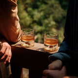 Two clear tumblers with amber liquid and visible dinosaur bone shards embedded inside, placed on a wooden railing in soft natural light with blurred green foliage background.