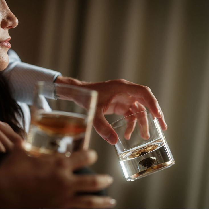 Close-up of two hands holding short clear tumblers with amber liquid and dark stone shards inside, set against a softly blurred beige background.