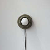 Minimalist wall sconce with an olive-green circular base and a central white globe bulb attached to a black cord hanging down on a light gray wall.