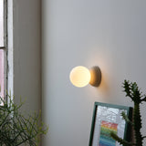 Glowing round white glass wall sconce with matte black base mounted on a light gray wall above a framed colorful abstract art and green spiky plants beside a window.
