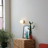 Minimalist wall with round frosted glass sconce above a natural wood cube table holding a green cactus in terracotta pot, turquoise hand sculpture, and framed artwork.