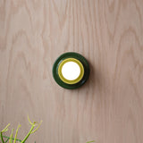 Round wall sconce with concentric dark green and yellow rings surrounding a bright central light, mounted on pale wood-paneled wall with small plant tips visible below.