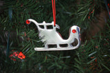 Silver sleigh-shaped Christmas ornament with red ruby-colored gems, hanging from a red string on green pine tree branches.