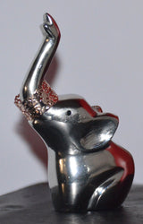 Small silver-finished elephant figurine with trunk raised, holding a delicate lace-patterned ring around its trunk, shown on a dark surface with plain background.