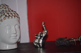 Silver elephant figurine with raised trunk and textured crown detail, placed between a white Buddha head sculpture and a miniature horse-drawn carriage against a red background.
