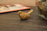 Small golden bird figurine with textured feather details on a dark wooden surface, with a glass jar of stones and an orange-edged book in the background.