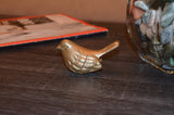 Small bronze bird figurine with textured feather detailing resting on a dark wooden surface, nearby glass vase filled with light-colored stones in soft focus.