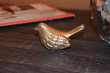 Small metallic gold bird figurine with detailed feather patterns resting on a dark wooden surface beside a glass container filled with stones.