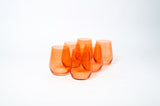 Six translucent orange stemless wine glasses grouped on a white background, varying slight in height and shape with smooth rounded edges.