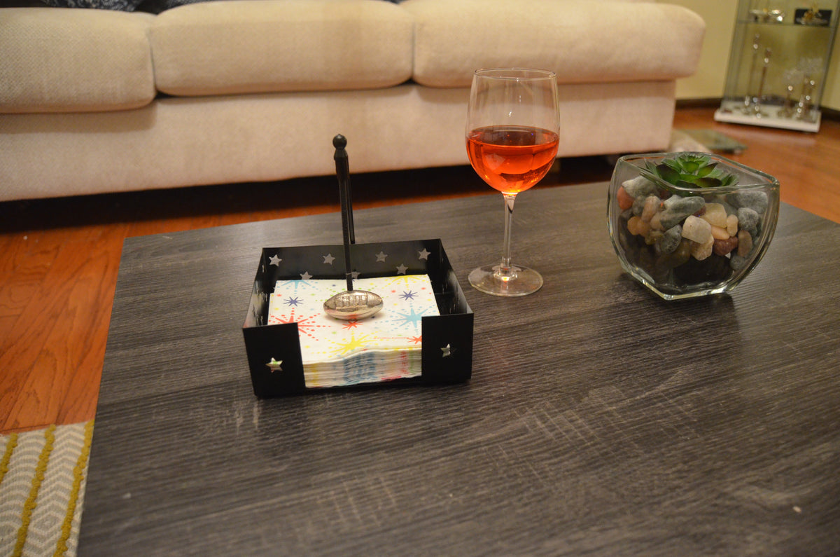 Black metal napkin holder with star-shaped cutouts holding colorful napkins, placed on a dark wood coffee table beside a glass of rosé wine and a succulent in a glass container.