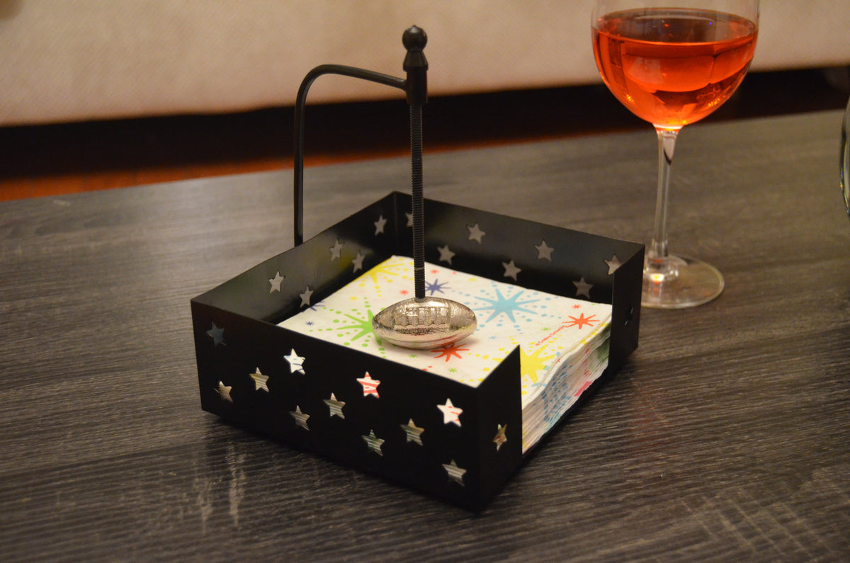 Black metal napkin holder with star-shaped cutouts and an American football-shaped silver handle, holding colorful starburst patterned napkins on a dark wood table.