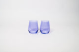 Two translucent stemless wine glasses in a soft periwinkle purple hue, placed side by side on a plain white surface.