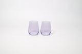Pair of stemless wine glasses in translucent lavender color with a smooth, curved silhouette, shown side by side on a white background.