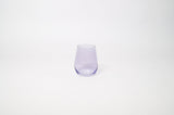 Stemless wine glass with a soft lavender tint holding white wine on a textured surface
