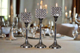 Set of three polished silver aluminum candle holders with square tops adorned by clear crystal circles, each holding a lit candle, arranged by height on a white tablecloth.