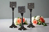 Set of three tall, slender aluminium candle holders with square tops adorned with clear crystal circles, displayed against a soft background with peach and white roses.