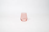 Tickle Me Pink stemless wine glass in soft pastel pink on a wooden surface with greenery in the background