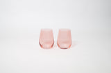 Two stemless wine glasses with a translucent pink tint, slightly tapered with curved bodies and smooth rims against a plain white background.