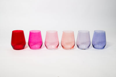 Six stemmed wine glasses in ombré colors ranging from pink to purple, arranged on a beige linen surface.