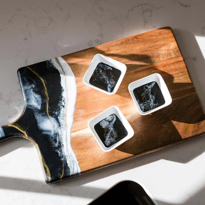 Wooden charcuterie board with black and white abstract resin design, holding four square items on a light surface.