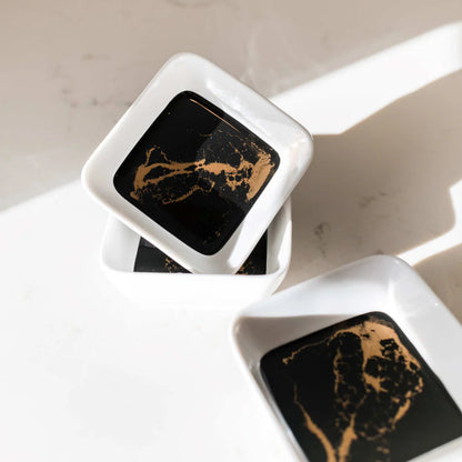 Two square black and gold resin ceramic dishes on a white surface