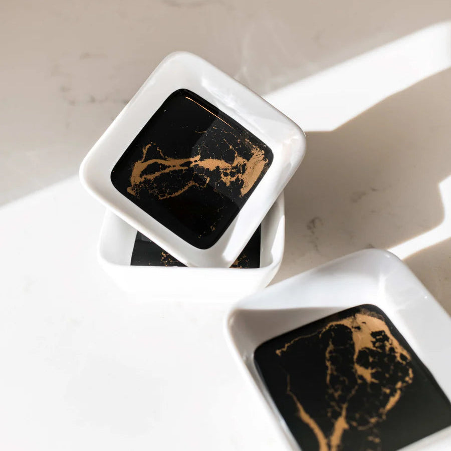 Two square black and gold resin ceramic dishes on a white surface
