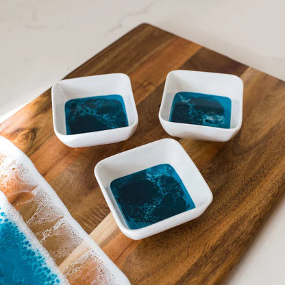 Three small resin white bowls with blue liquid on a wooden cutting board.