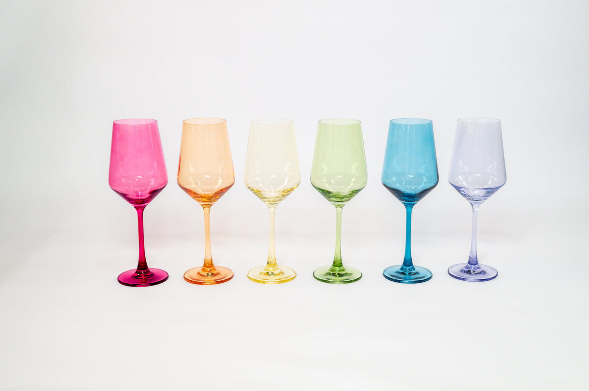 Six pastel-toned wine glasses, arranged in a row, colored pink, orange, yellow, green, blue, and lavender, against a plain white background.