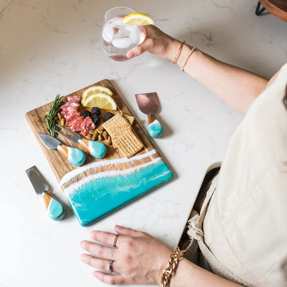 Person holding a glass of water with lemon slices next to a resin charcuterie board on a marble surface.