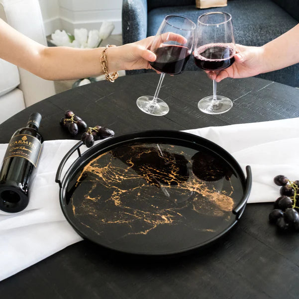 Two people toasting with red wine glasses on a table with a resin tray and bottle of wine.