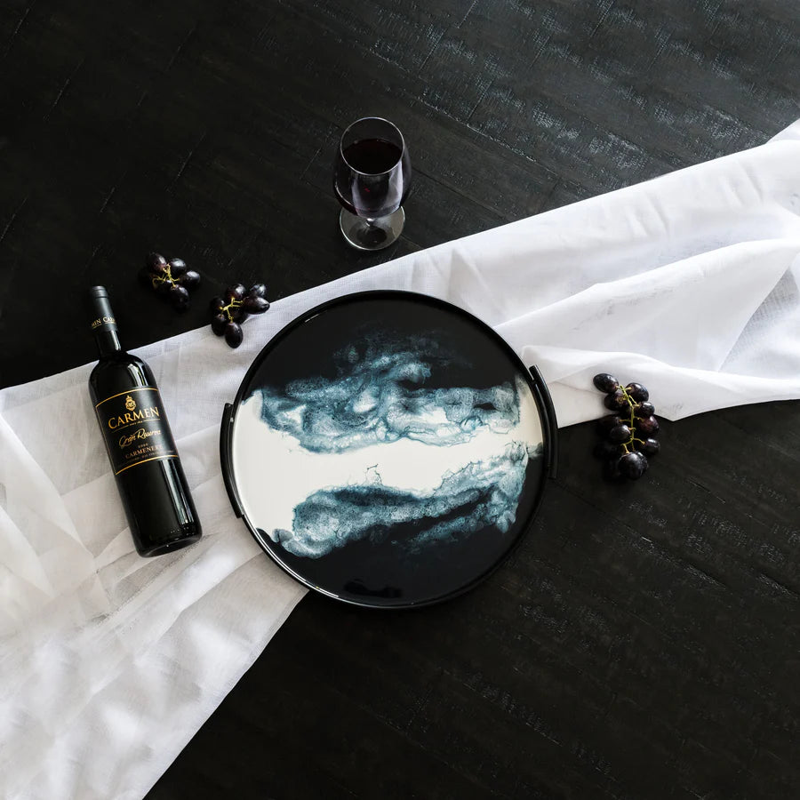 Round decorative resin tray with artistic design on a black table with a bottle of wine and grapes.